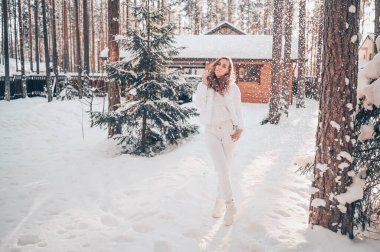 Kar yağışı altında güneşli soğuk bir portre. Beyaz, kalın bir ceketle, kır evinin arka bahçesinde kar kaplı çam ağaçlarıyla kaplı yeşil çam ağaçlarının arka bahçesinde.