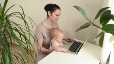 Genç anne karantina sırasında dizüstü bilgisayarla evde çalışıyor. Kucağında küçük şirin bir bebek var. Merkez ofis, ebeveynlik. Uzaktan çalışma, uzaktan eğitim. Video konferansı sanal olay.
