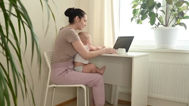 Νεαρή μητέρα εργάζεται μελετώντας από το σπίτι με φορητό υπολογιστή κατά τη διάρκεια της καραντίνας, λίγο χαριτωμένο μωρό στην αγκαλιά. Γραφείο στο σπίτι, γονιός. Απομακρυσμένη δουλειά, εξ αποστάσεως εκπαίδευση. Εικονική εκδήλωση τηλεδιάσκεψης. — Αρχείο Βίντεο