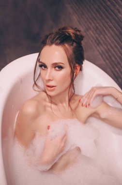 Makyajlı ve saç stili olan güzel genç bir kadın köpük dolu eski bir küvette banyo yaparken rahatlıyor. Spa ve güzellik salonu konsepti, vücut ve cilt bakımı.