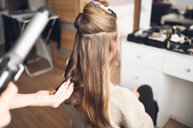 Saç stilisti, güzel bir genç kadını etkinliğe hazırlar, kuaför salonunda müşterileri için saç maşası ile saç stili yapar. Uzun, açık kahverengi, doğal saçlar..