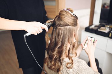 Saç stilisti, güzel bir genç kadını etkinliğe hazırlar, kuaför salonunda müşterileri için saç maşası ile saç stili yapar. Uzun, açık kahverengi, doğal saçlar..