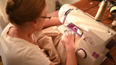 Kendinden emin yaşlı kadın terzi beyaz gömlek ve gözlükler dikiş makinesinin önünde oturuyor, bej kumaş kullanarak evde giysiler üzerinde çalışıyor. Emekli insanlar, yaş, iş hayatı kavramı.