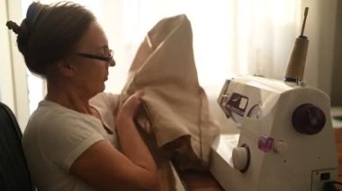 Kendinden emin yaşlı kadın terzi beyaz gömlek ve gözlükler dikiş makinesinin önünde oturuyor, bej kumaş kullanarak evde giysiler üzerinde çalışıyor. Emekli insanlar, yaş, iş hayatı kavramı.