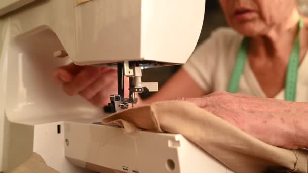 Confiada anciana mayor costurera camisa blanca y gafas sentadas en frente de la máquina de coser, trabajando en la ropa en casa usando tela beige. Personas jubiladas, edad, concepto de ocupación laboral. — Vídeos de Stock