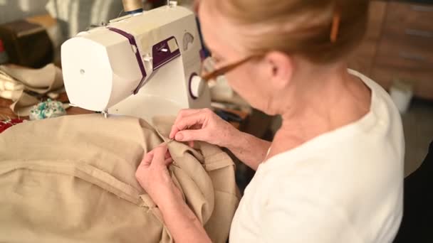 Magabiztos idős nő varrónő fehér ing és szemüveg ül a varrógép előtt, dolgozik a ruhák otthon bézs szövet. Nyugdíjasok, életkor, foglalkozás fogalma. — Stock videók
