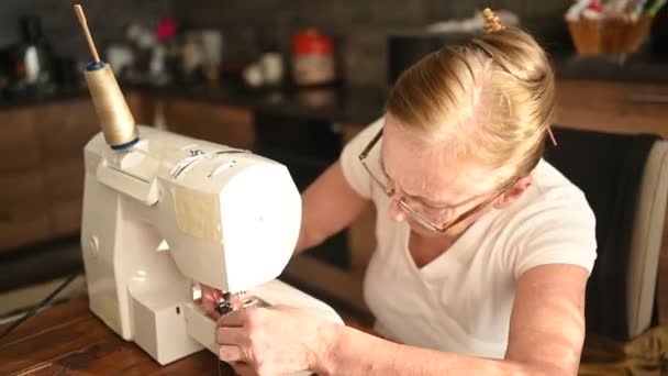 Confiada anciana mayor costurera camisa blanca y gafas sentadas en frente de la máquina de coser, trabajando en la ropa en casa usando tela beige. Personas jubiladas, edad, concepto de ocupación laboral. — Vídeos de Stock