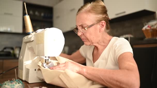 Confident senior elderly woman seamstress white shirt and glasses sitting in front of sewing machine, working on clothes at home using beige fabric. Retired people, age, job occupation concept. — Stock Video