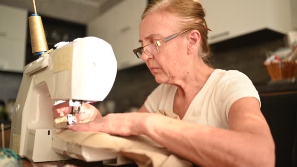 Fiduciosa anziana donna anziana cucitrice camicia bianca e occhiali seduto di fronte alla macchina da cucire, lavorando sui vestiti a casa con tessuto beige. Pensionati, età, concetto di occupazione. — Video Stock