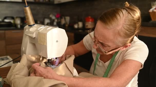 विश्वस्त वरिष्ठ बुजुर्ग महिला seamstress सफेद शर्ट और चश्मा सिलाई मशीन के सामने बैठे, बेज कपड़े का उपयोग करके घर पर कपड़े पर काम कर रहे हैं। सेवानिवृत्त लोग, उम्र, नौकरी व्यवसाय अवधारणा . — स्टॉक वीडियो