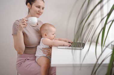 Genç anne karantina sırasında dizüstü bilgisayarla evde çalışıyor. Kucağında küçük şirin bir bebek var. Merkez ofis, ebeveynlik. Uzaktan çalışma, uzaktan eğitim. Video konferansı sanal olay