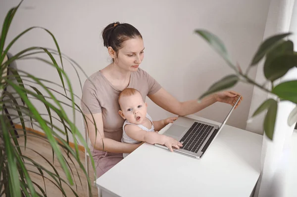 Young mother work studying from home with laptop computer during quarantine, little cute toddler baby on lap. Home office, parenthood. Remote work, distance education. Video conference virtual event