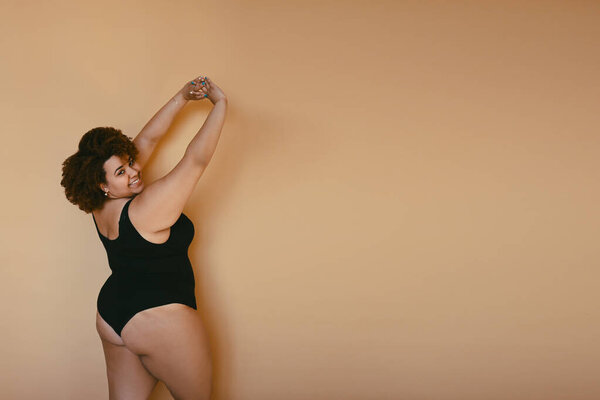Beautiful curvy oversize African black woman afro hair posing in black bodysuit on beige brown background isolated, body imperfection, body acceptance, body positive and diversity concept. Copyspace.