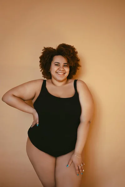Mooie ronding oversized Afrikaanse zwarte vrouw afro haar poseren in zwart bodysuit op beige bruine achtergrond geïsoleerd, lichaam imperfectie, aanvaarding van het lichaam, lichaam positief en diversiteit concept. Kopieerruimte. Stockfoto