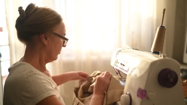 Kendinden emin yaşlı kadın terzi beyaz gömlek ve gözlükler dikiş makinesinin önünde oturuyor, bej kumaş kullanarak evde giysiler üzerinde çalışıyor. Emekli insanlar, yaş, iş hayatı kavramı. — Stok video