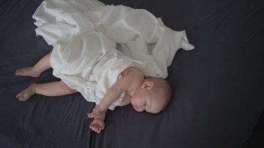 Beyaz çarşafla örtülmüş, gri yatakta uyuyan sevimli yeni doğmuş bebek. 