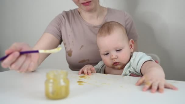 Pequeño bebé divertido recién nacido aprendiendo a comer puré de verduras o frutas de frasco de vidrio con cuchara. Joven madre ayudando a su pequeño hijo a comer primera comida. — Vídeo de stock