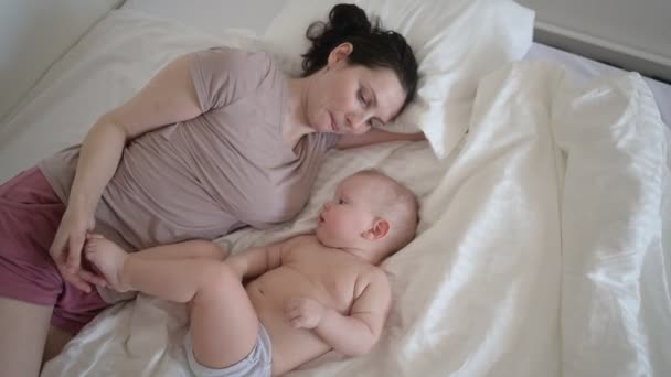 Mutlu genç bir anne, tatlı bir bebekle yatakta yatıyor, onu kollarına alıyor, sarılıyor ve sabahın erken saatlerinde oynuyor. Sağlıklı çocuk, hastane kavramı ve mutlu annelik. Çocuk odası. — Stok video