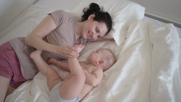 Szczęśliwa młoda matka leżąca z słodkim malutkim chłopczykiem na łóżku, trzymająca go na rękach, przytulająca i bawiąca się wcześnie rano. Zdrowe dziecko, koncepcja szpitala i szczęśliwe macierzyństwo. Przedszkole — Wideo stockowe