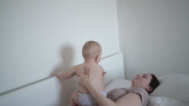 Mutlu genç bir anne, tatlı bir bebekle yatakta yatıyor, onu kollarına alıyor, sarılıyor ve sabahın erken saatlerinde oynuyor. Sağlıklı çocuk, hastane kavramı ve mutlu annelik. Çocuk odası.