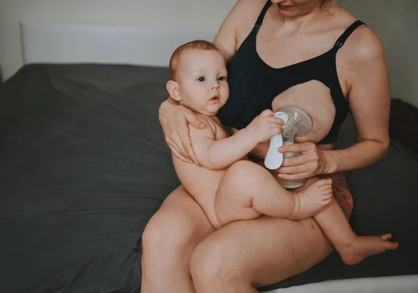 Young mother with newborn cute infant naked baby boy, holding him on arms, using breast pump hugging and breastfeeds with breast milk. Healthy child, concept of hospital and happy motherhood. Nursery. — Stock Photo, Image