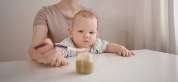 Pequeño bebé divertido recién nacido aprendiendo a comer puré de verduras o frutas de frasco de vidrio con cuchara. Joven madre ayudando a su pequeño hijo a comer primera comida. —  Fotos de Stock