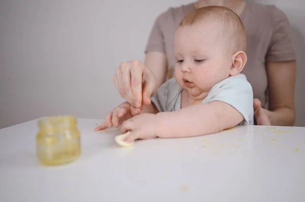 Pequeño bebé divertido recién nacido aprendiendo a comer puré de verduras o frutas de frasco de vidrio con cuchara. Joven madre ayudando a su pequeño hijo a comer primera comida. —  Fotos de Stock