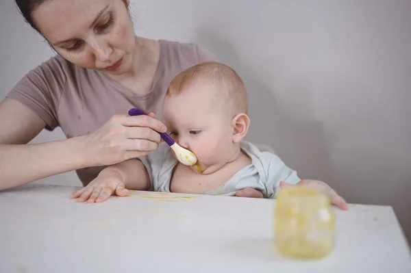 Menino bebê engraçado recém-nascido aprendendo a comer purê de vegetais ou frutas de jarra de vidro com colher. Jovem mãe ajudando pequeno filho comer primeira comida. — Fotografia de Stock