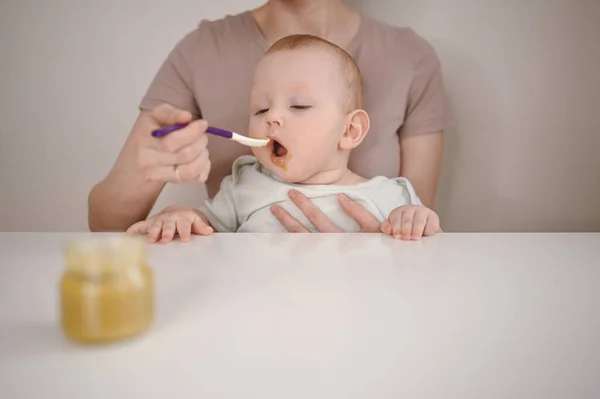 Pequeño bebé divertido recién nacido aprendiendo a comer puré de verduras o frutas de frasco de vidrio con cuchara. Joven madre ayudando a su pequeño hijo a comer primera comida. —  Fotos de Stock