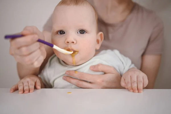 Pequeño bebé divertido recién nacido aprendiendo a comer puré de verduras o frutas de frasco de vidrio con cuchara. Joven madre ayudando a su pequeño hijo a comer primera comida. —  Fotos de Stock