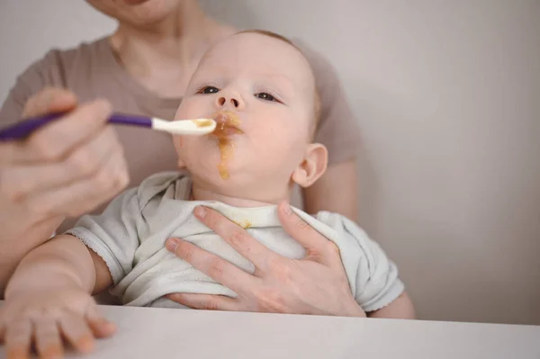 Pequeño bebé divertido recién nacido aprendiendo a comer puré de verduras o frutas de frasco de vidrio con cuchara. Joven madre ayudando a su pequeño hijo a comer primera comida. —  Fotos de Stock