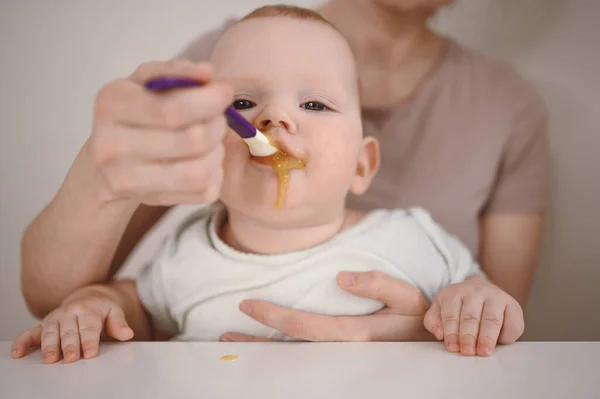 Pequeño bebé divertido recién nacido aprendiendo a comer puré de verduras o frutas de frasco de vidrio con cuchara. Joven madre ayudando a su pequeño hijo a comer primera comida. —  Fotos de Stock