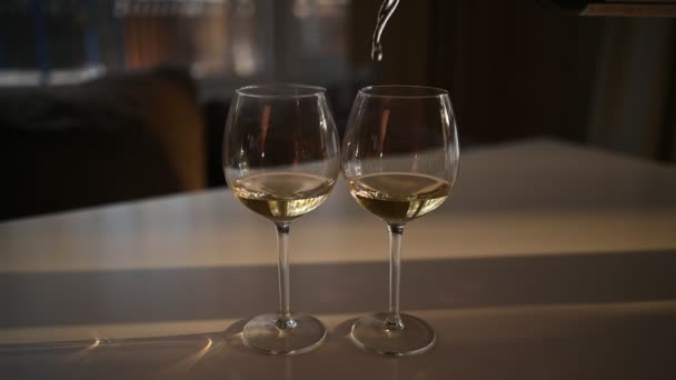 Nalije bílé suché víno do jedné ze dvou sklenic stojící na bílém stole. Rozostřené rozmazané pozadí. Zavřít zpomalené video procesu míchání vína v poháru se slunečním světlem — Stock video