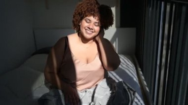Güzel kıvrımlı, iri yapılı, Afro saçlı Afrikalı bir kadın. Gri battaniye örtülü, yatak odalı, iç tasarımı var. Güneş ışığı. Vücut kusuru, beden kabulü, vücut pozitifliği ve çeşitlilik kavramı