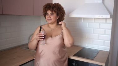 Güzel kıvrımlı, iri yapılı, Afrikalı, afro saçlı bir kadın modern İskandinav tarzı mutfak iç tasarımında kırmızı şarap içiyor. Vücut kusuru, beden kabulü, vücut pozitifliği ve çeşitlilik kavramı