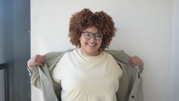 Mooie vrolijke glimlach ronding plus size Afrikaanse zwarte vrouw afro haar poseren in beige t-shirt, jeans, stijlvolle bril op zonnig balkon. Lichaamsimperfectie, acceptatie lichaam positief diversiteitsconcept — Stockvideo