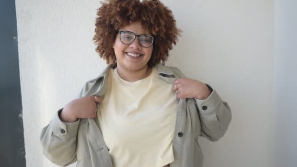 Hermosa feliz sonrisa curvas más el tamaño Africano negro mujer afro pelo posando en camiseta beige, jeans, gafas con estilo en el balcón soleado. Imperfección corporal, aceptación cuerpo concepto de diversidad positiva — Vídeos de Stock