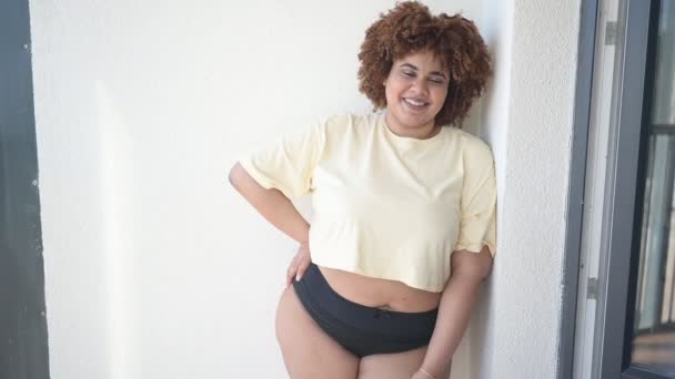 Mooie vrolijke ronding plus size Afrikaanse zwarte vrouw afro haar poseren in beige t-shirt en zwart ondergoed op zonnig balkon. Lichaamsimperfectie, lichaamsacceptatie, lichaamspositief en diversiteitsconcept — Stockvideo