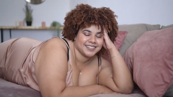 Mooie ronding plus size Afrikaanse zwarte vrouw afro haar liggend op bed in zijden poeder roze jurk gezellige slaapkamer interieur. Lichaamsimperfectie, lichaamsacceptatie, lichaamspositief en diversiteitsconcept — Stockvideo