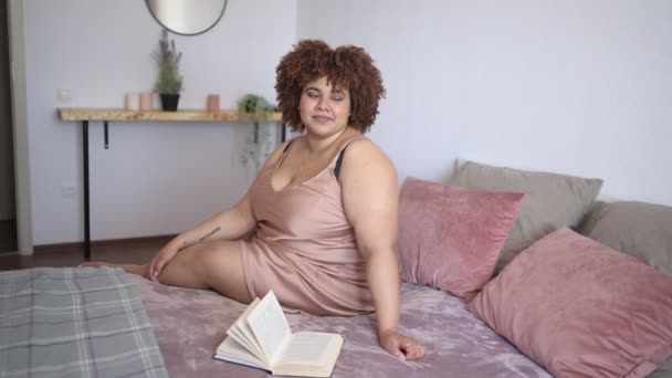 Mooie ronding plus size Afrikaanse zwarte vrouw afro haar liggend op bed in zijden poeder roze jurk gezellige slaapkamer interieur. Lichaamsimperfectie, lichaamsacceptatie, lichaamspositief en diversiteitsconcept — Stockvideo