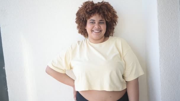 Mooie vrolijke ronding plus size Afrikaanse zwarte vrouw afro haar poseren in beige t-shirt en zwart ondergoed op zonnig balkon. Lichaamsimperfectie, lichaamsacceptatie, lichaamspositief en diversiteitsconcept — Stockvideo