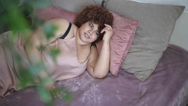 Hermoso curvas más tamaño africano negro mujer afro pelo acostado en la cama en polvo de seda vestido rosa acogedor dormitorio diseño interior. Imperfección corporal, aceptación corporal, concepto de cuerpo positivo y diversidad — Vídeos de Stock