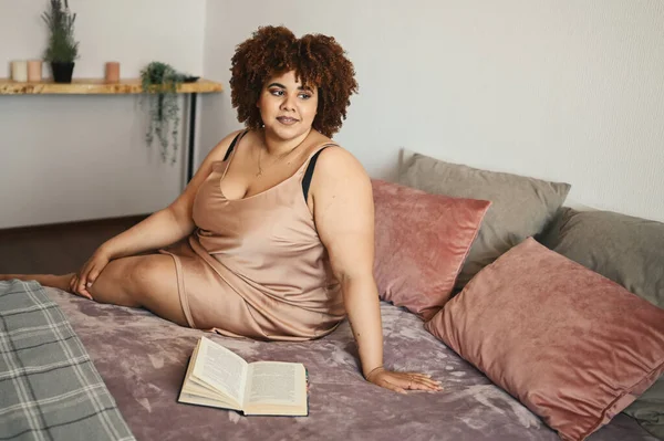 Linda curvilínea plus size Africano preto mulher afro cabelo deitado na cama em pó de seda rosa vestido aconchegante quarto design de interiores. Imperfeição corporal, aceitação corporal, corpo positivo e conceito de diversidade — Fotografia de Stock