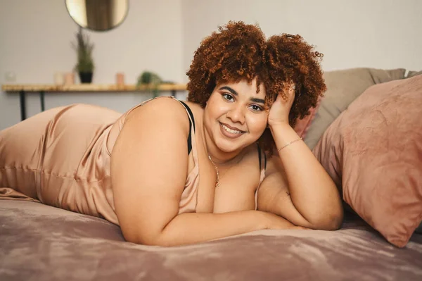 Beautiful curvy plus size African black woman afro hair lying on bed in silk powder pink dress cozy bedroom interior design. Body imperfection, body acceptance, body positive and diversity concept