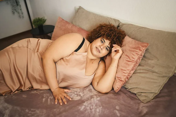 Indah melengkung ditambah ukuran Afrika hitam wanita afro rambut berbaring di tempat tidur dalam bubuk sutra merah muda gaun nyaman desain interior. Ketidaksempurnaan tubuh, penerimaan tubuh, tubuh konsep positif dan keragaman — Stok Foto