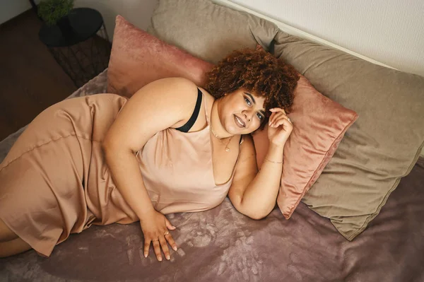 โค้งที่สวยงามบวกขนาดแอฟริกันผู้หญิงผมสีดํา afro นอนบนเตียงในผงผ้าไหมชุดสีชมพูการออกแบบตกแต่งภายในห้องนอนที่อบอุ่น ความไม่สมบูรณ์ของร่างกาย การยอมรับร่างกาย ความคิดเชิงบวกและความหลากหลายของร่างกาย — ภาพถ่ายสต็อก