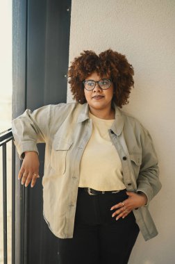 Güzel, mutlu, gülümseyen kıvrımlı, kahverengi saçlı, bej tişörtlü, kot pantolonlu, güneşli balkonda şık gözlüklü bir kadın. Vücut kusuru, kabullenme vücut pozitif çeşitlilik kavramı