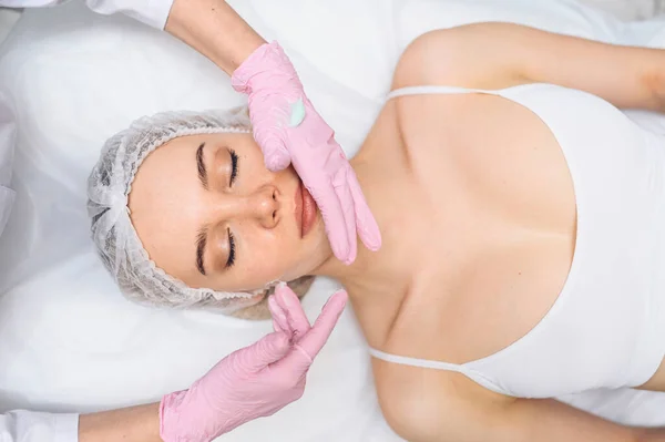 Wanita cantik yang bahagia menerima perawatan spa. Kosmetologis di salon kecantikan dengan sarung tangan merah muda menerapkan masker krim pelembab. Keindahan wajah. Sempurna segar bersih kulit. Konsep perawatan anak muda dan kulit. — Stok Foto