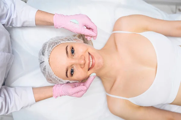 Wanita cantik yang bahagia menerima perawatan spa. Kosmetologis di salon kecantikan dengan sarung tangan merah muda menerapkan masker krim pelembab. Keindahan wajah. Sempurna segar bersih kulit. Konsep perawatan anak muda dan kulit. — Stok Foto