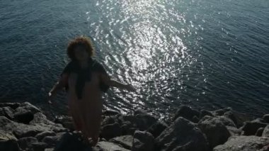 Mutlu ve boyut Afrikalı siyah kadın afro saçlar gün batımında deniz kenarında tatilin tadını çıkarıyor güneşli hava, kayalar, mavi okyanus suyu. Vücut kusuru, kabullenme vücut pozitif çeşitlilik kavramı